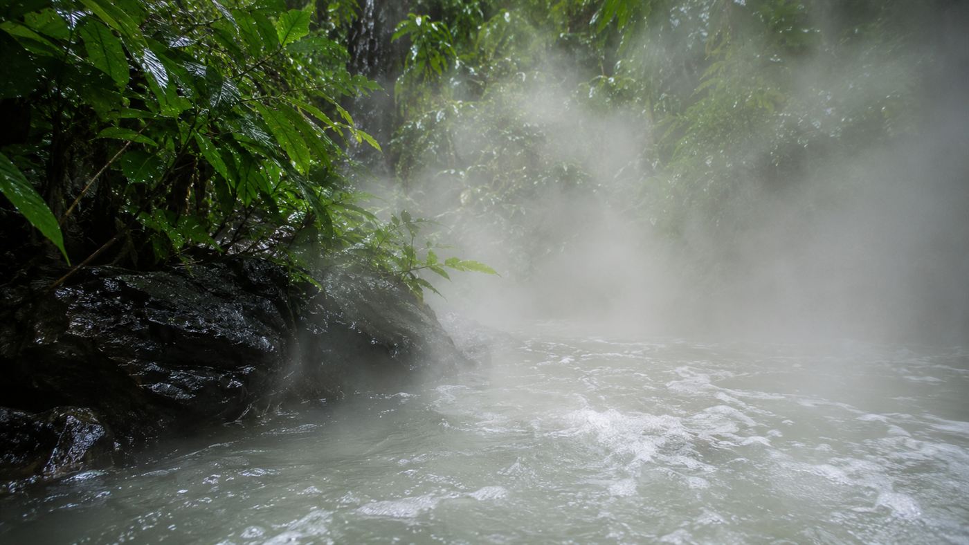 Close view of the surface of a hot river giving off thick steam, with dark rocks at the edge and green rainforest pressing in on both banks