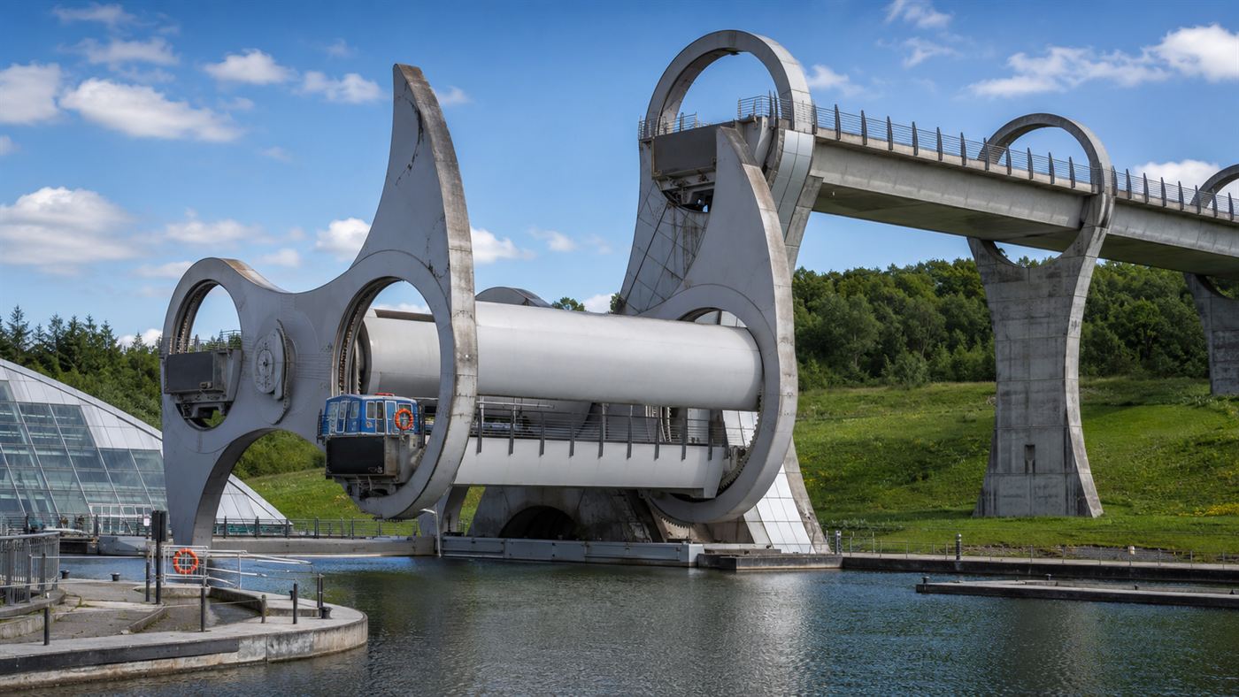 A small boat sitting inside a water-filled gondola of the Falkirk Wheel as the great arm lifts it up between the canal basin and a high aqueduct