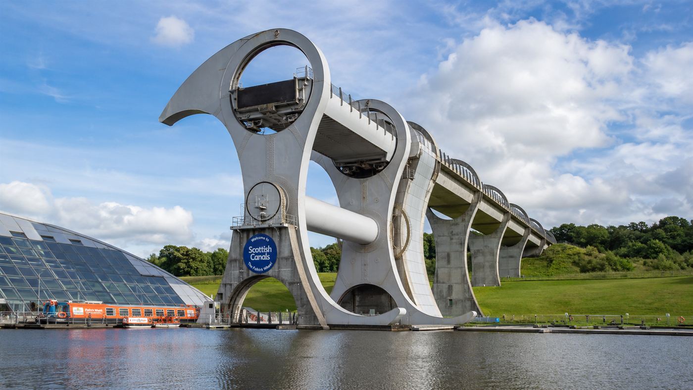 The Falkirk Wheel, a huge modern rotating boat lift with two curved steel arms and water-filled gondolas, beside a canal basin in Scotland under a bright sky