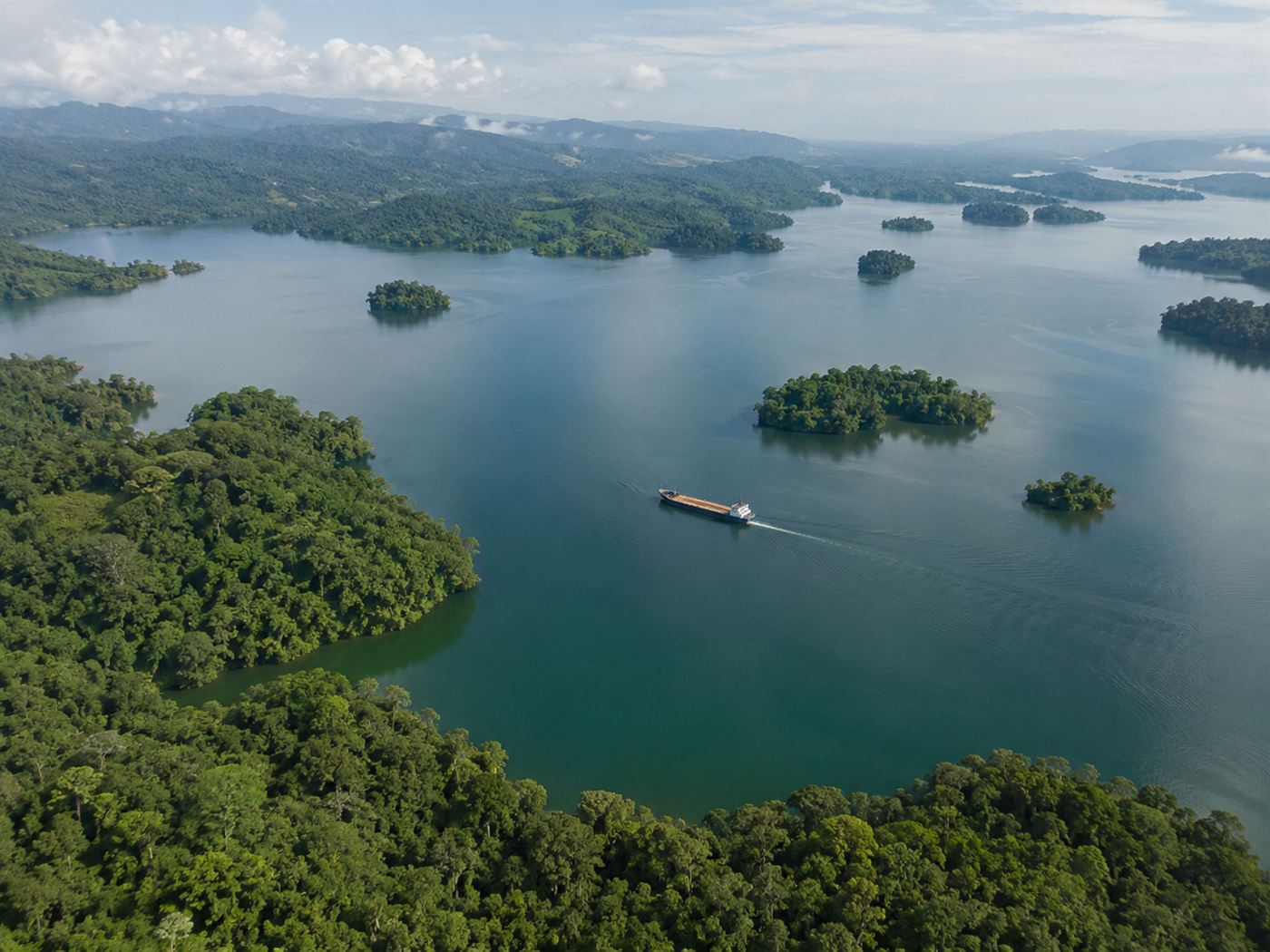 An aerial view of a large tropical freshwater lake surrounded by dense green rainforest, with a cargo ship crossing it