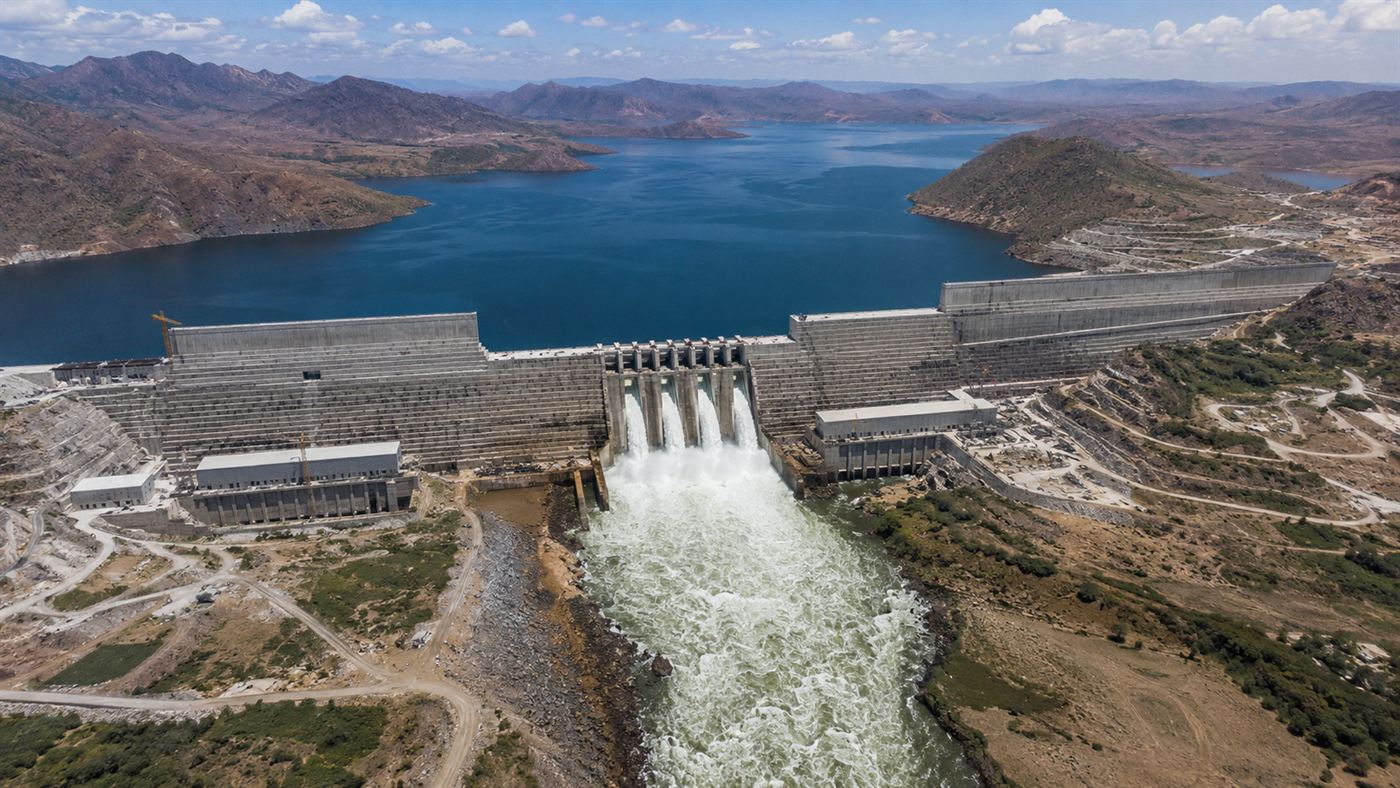 An enormous concrete hydroelectric dam spanning a wide river gorge in the Ethiopian highlands, with white water rushing from the spillways and a large reservoir behind it