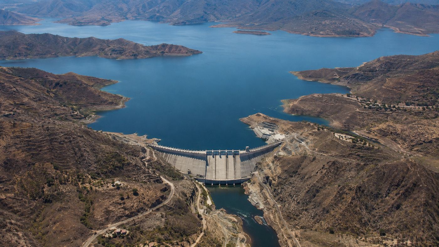 An aerial view of the vast blue reservoir held back by a large hydroelectric dam in a dry highland landscape