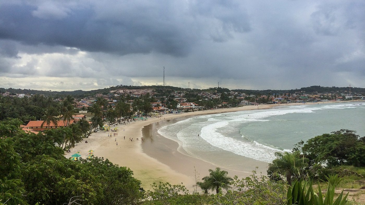 Praia de areia em curva com ondas brancas, casas e coqueiros, vista do alto sob céu nublado