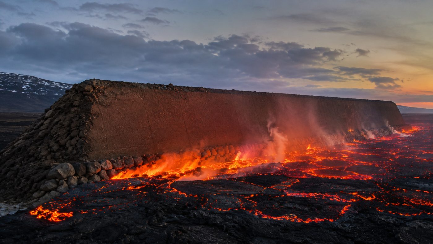 A massive earth and rock barrier wall built across an Icelandic lava field with glowing molten lava piling up against its base at dusk