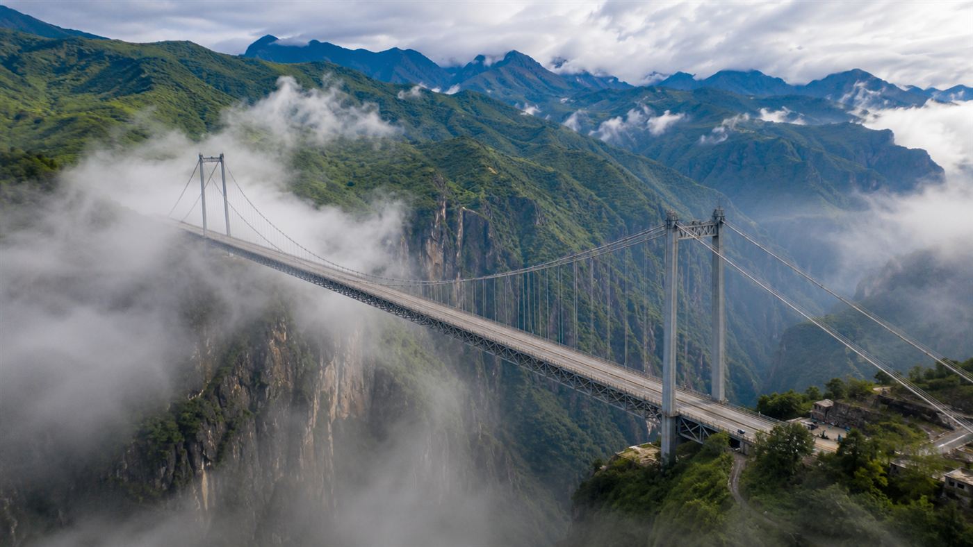 An aerial view of a steel suspension bridge spanning a wide misty gorge in mountainous terrain, with green peaks rolling into the distance