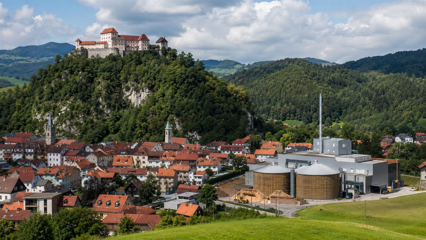 A small Austrian town with red roofs and a castle on a hill, surrounded by green forested hills, with a biomass heating plant and wood-chip silos at its edge