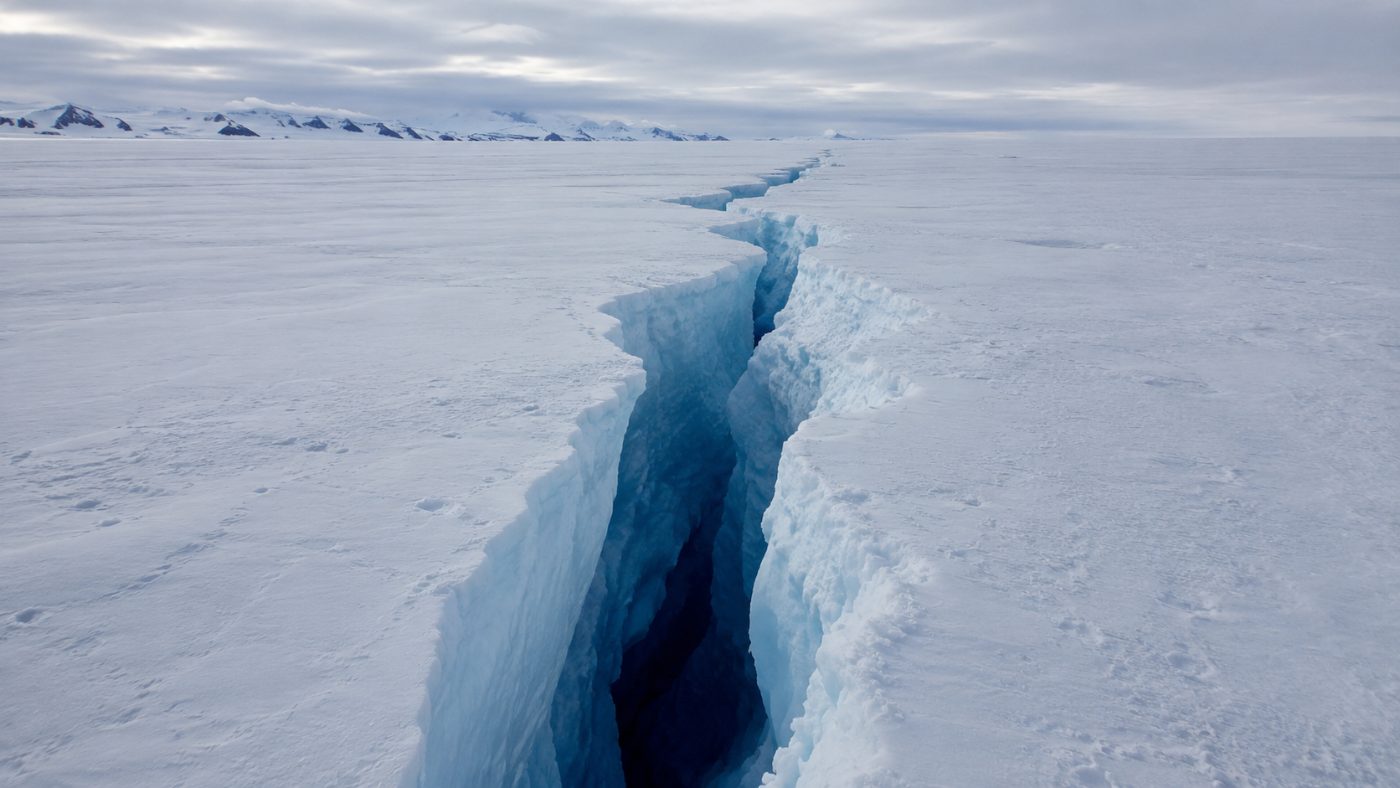 A deep jagged crack splitting across a vast flat white Antarctic ice shelf under an overcast sky