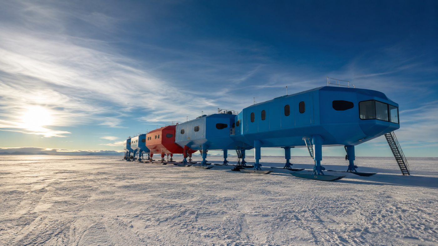A row of blue and red modular pods on hydraulic legs and skis standing on the flat white Antarctic ice shelf