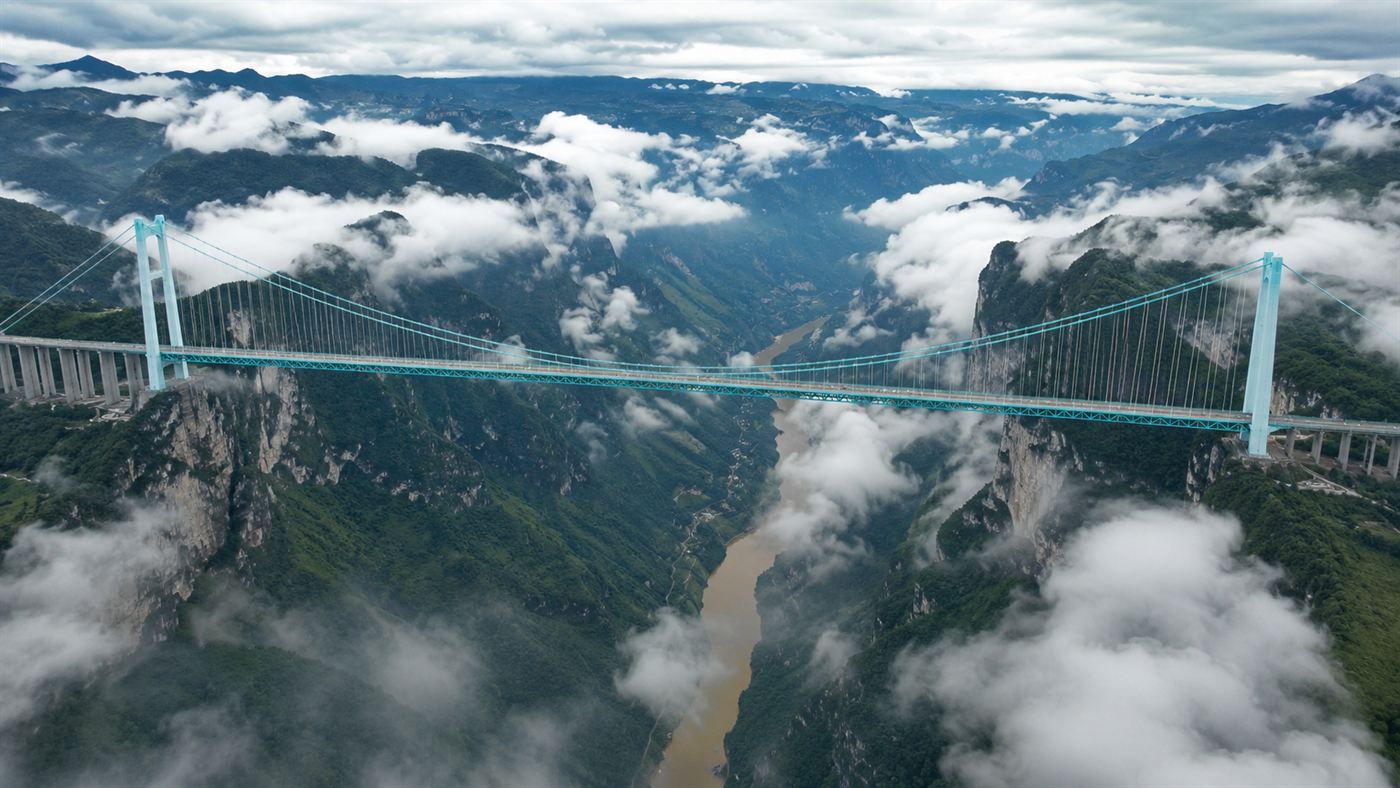 An enormous tall suspension bridge spanning a vast deep misty river canyon in mountainous Guizhou China, with clouds drifting below the bridge deck