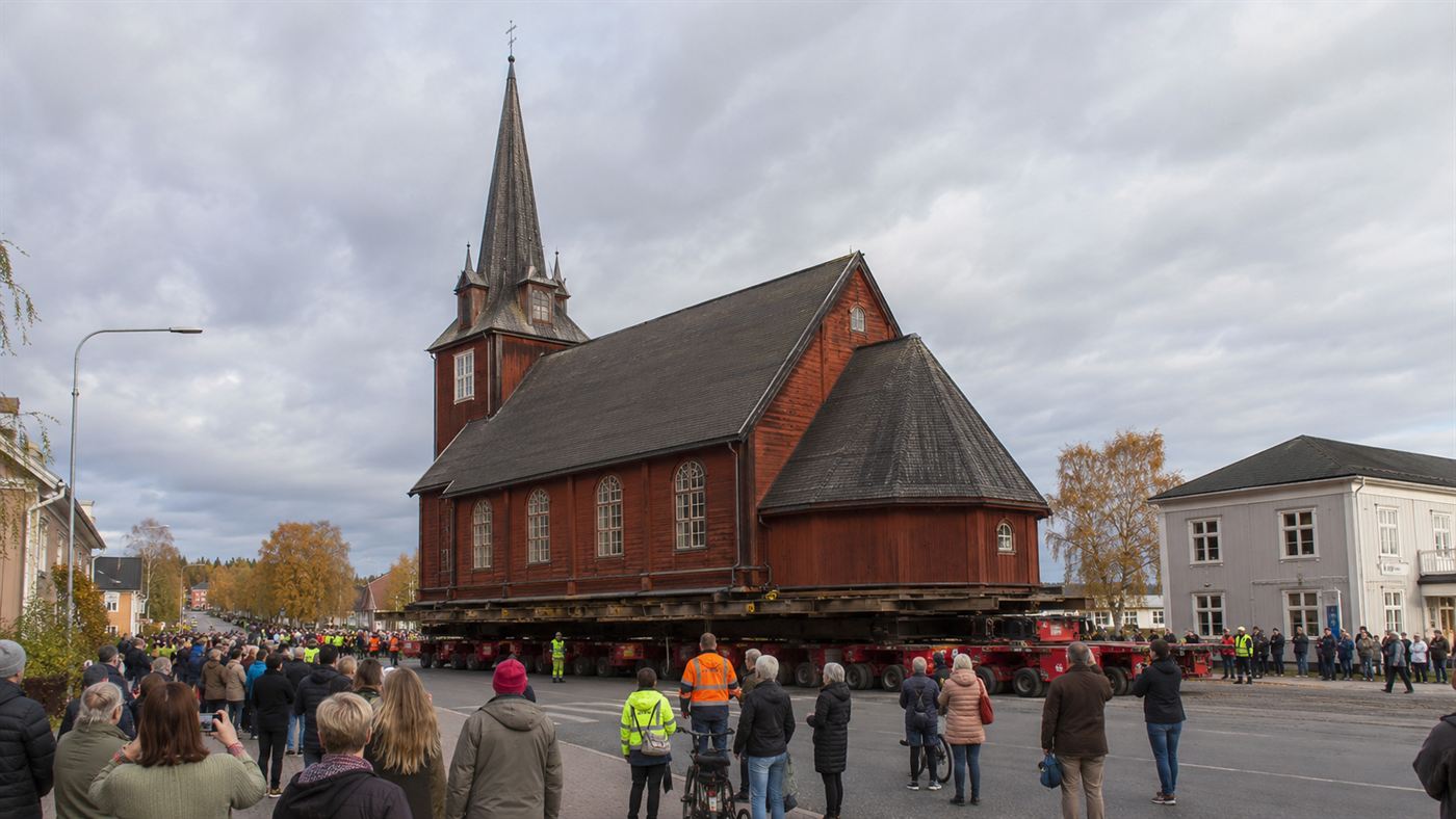 A large dark-red wooden church being moved whole on giant trailers along a road through an Arctic Swedish town