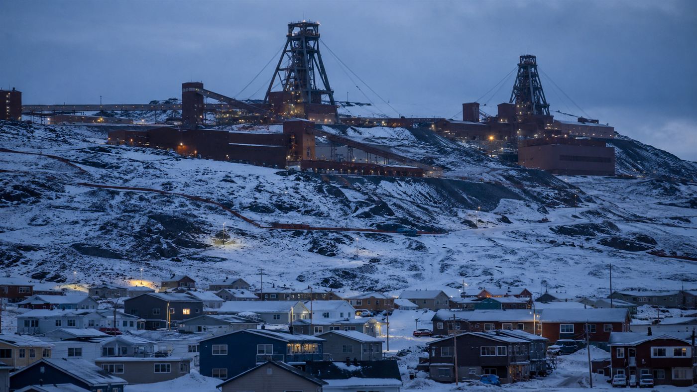 An Arctic mining town under snow with the huge industrial headframe and processing buildings of an iron ore mine looming on the hillside above the houses