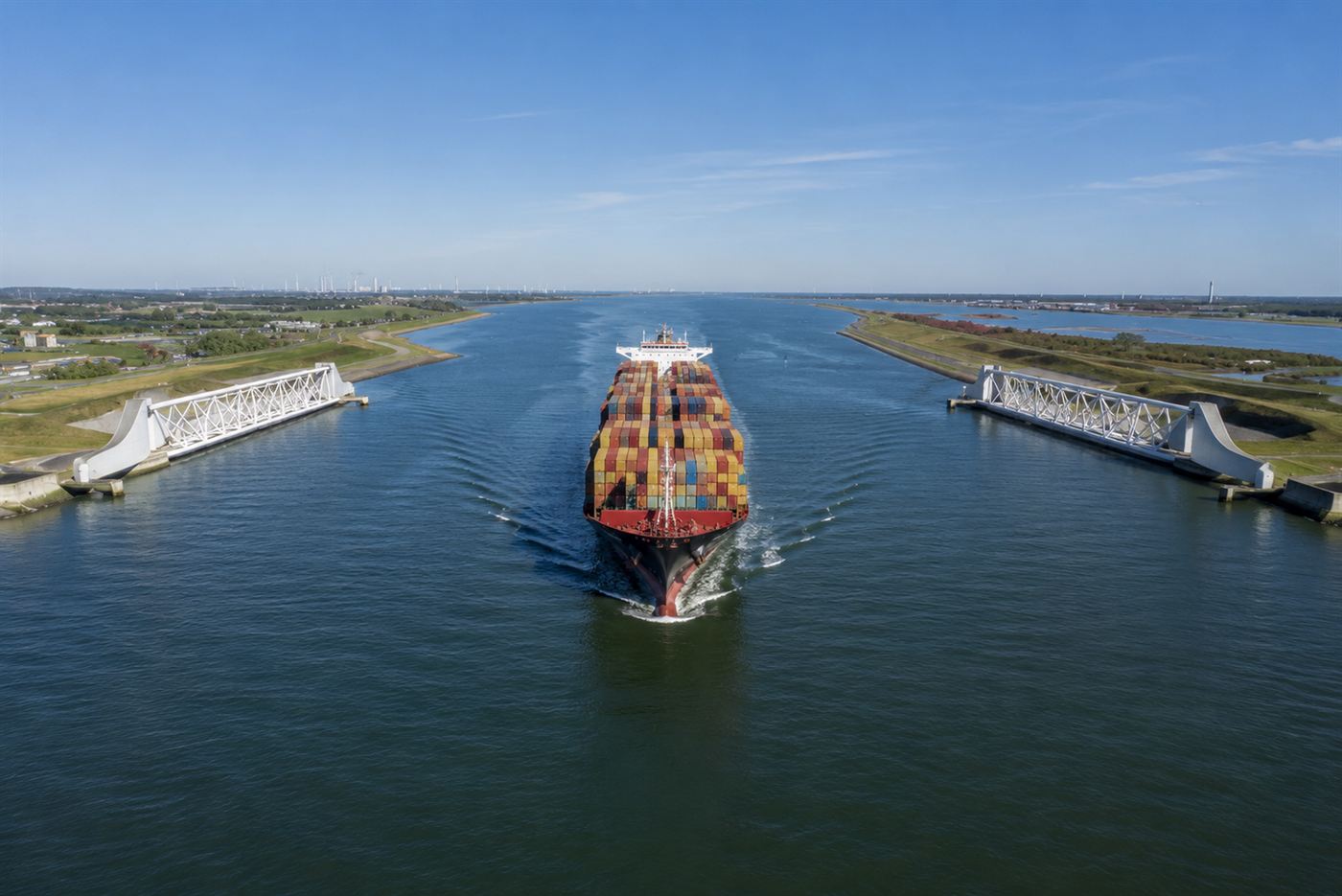 A large container ship sailing through the open Maeslant barrier between its two parked arms on a calm clear day