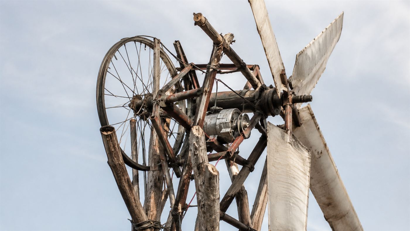 Close view of a homemade wind turbine built from a bicycle wheel and frame, a dynamo and flattened plastic blades mounted on a tower of wooden poles