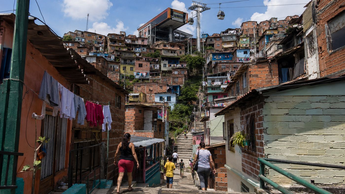 A hillside neighbourhood of densely packed colourful brick houses climbing a steep slope in Medellin, with a modern cable car station and gondolas above the rooftops