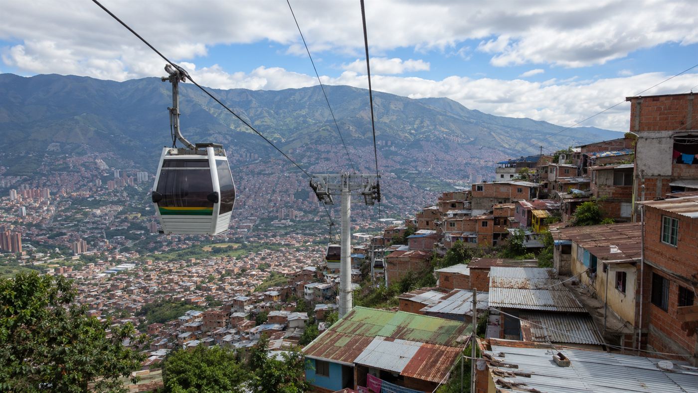 A cable car gondola rising over a dense hillside barrio of brick houses in Medellin