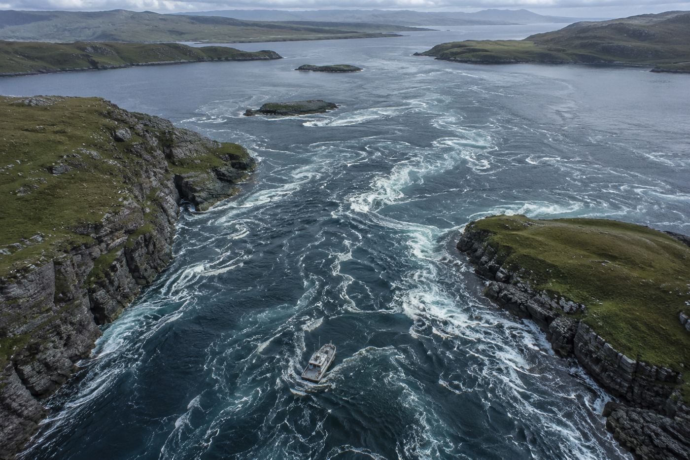 Aerial view of powerful tidal currents swirling through a narrow channel between rugged Scottish islands