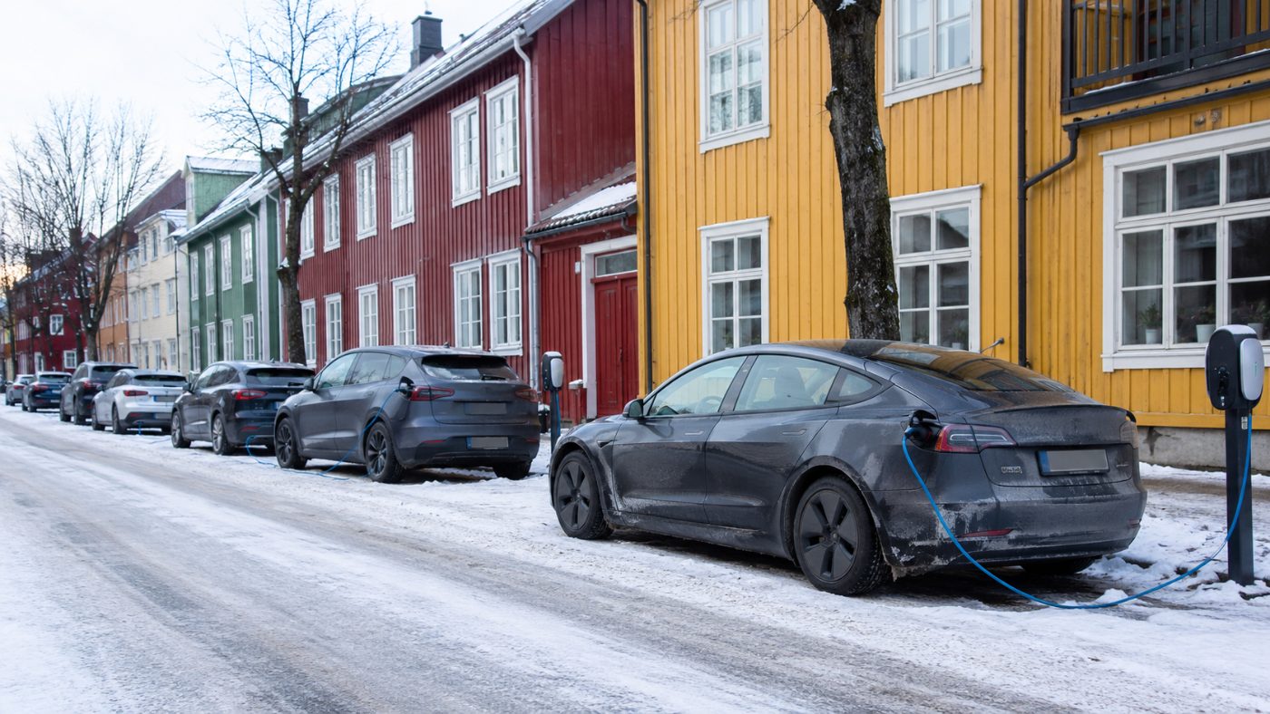 A snowy street in a Norwegian city lined with electric cars plugged into charging points beside colourful wooden buildings