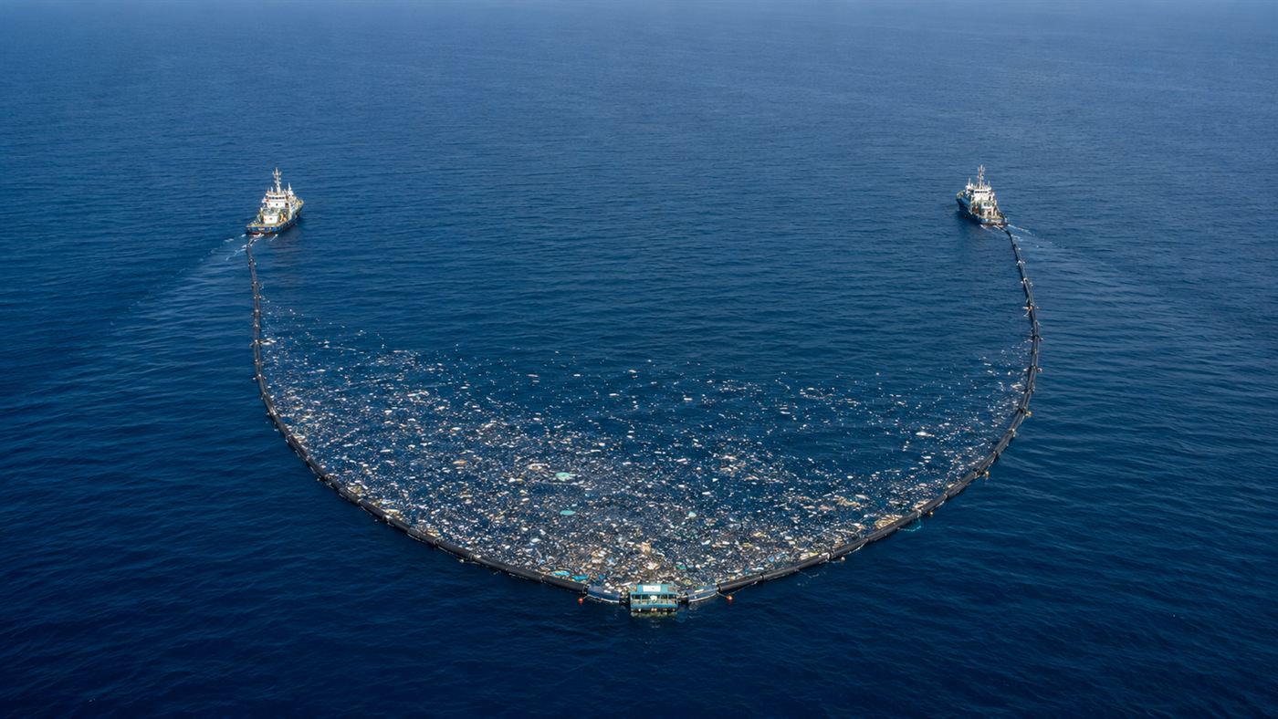 A long U-shaped floating barrier towed between two ships across the open ocean, gathering floating plastic into a retention zone