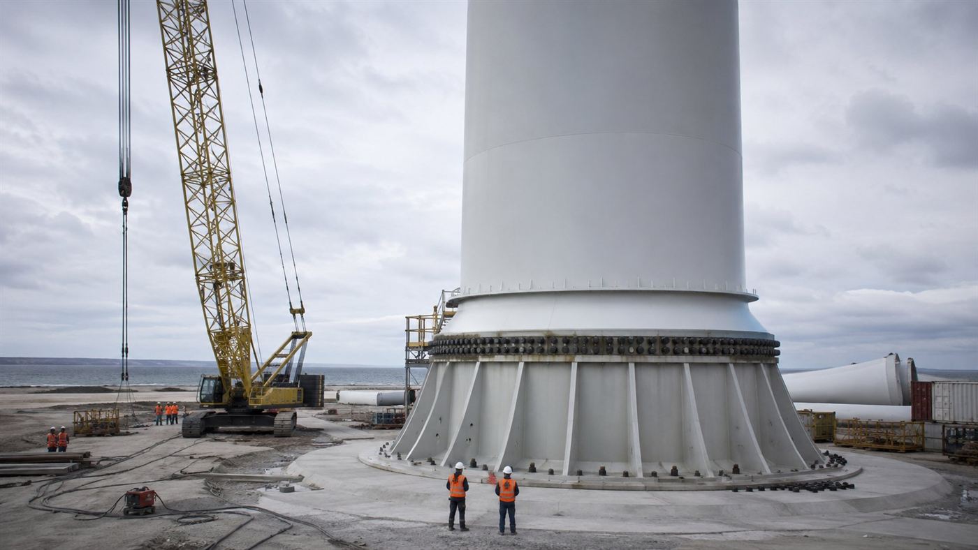 Engineers and a tall installation crane working on the base of a giant wind turbine tower at a coastal testing site under an overcast sky