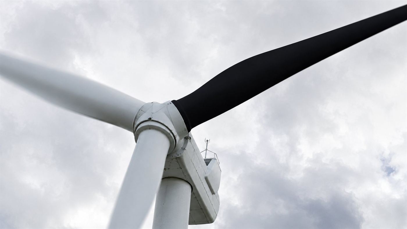 Close-up looking up at a wind turbine hub where one of the three blades is painted black and the other two are white against a bright sky