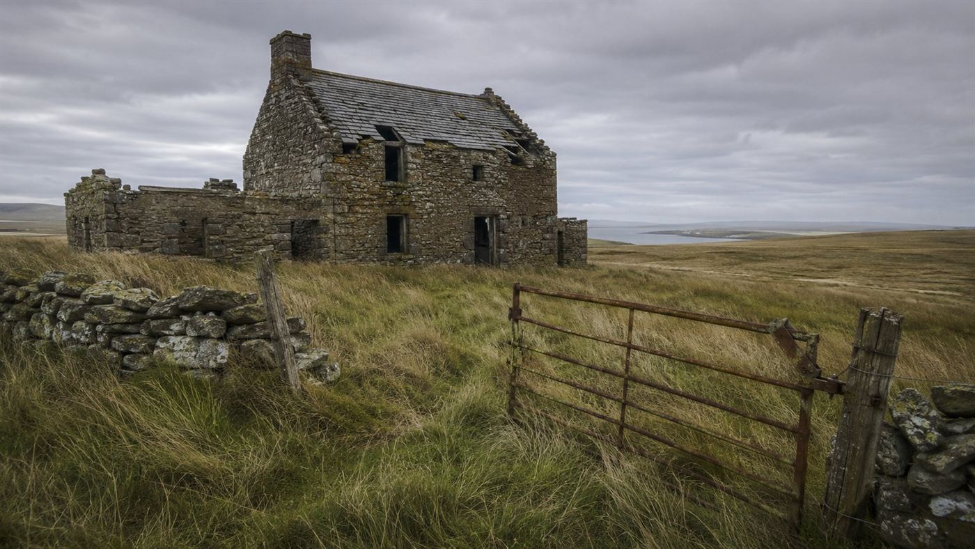 An abandoned stone farmhouse with empty windows on a remote treeless Scottish island under a grey overcast sky