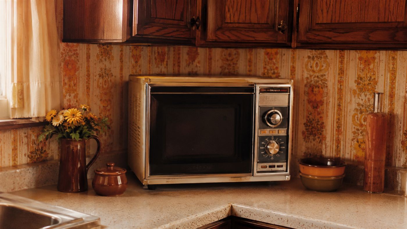 A 1970s countertop microwave oven in a retro home kitchen with wood cabinets and patterned wallpaper