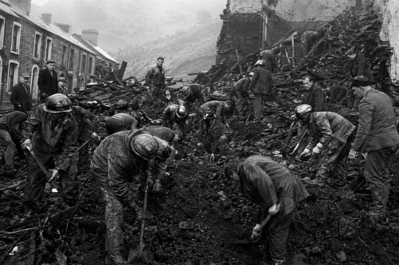Rescue workers digging through coal debris and mud in a destroyed building site in a Welsh village in 1966, many workers with shovels searching rubble in urgent grim scene, Aberfan coal tip collapse aftermath