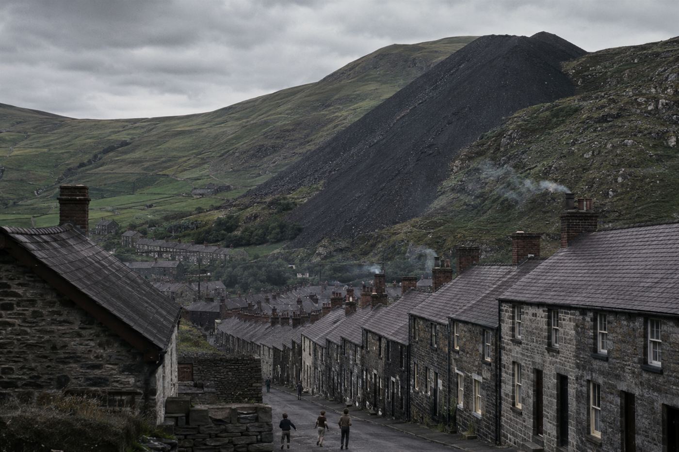 A Welsh valley mining village with a black coal waste slag tip looming on a steep hillside above rows of stone cottages, overcast grey sky, Aberfan 1966 disaster