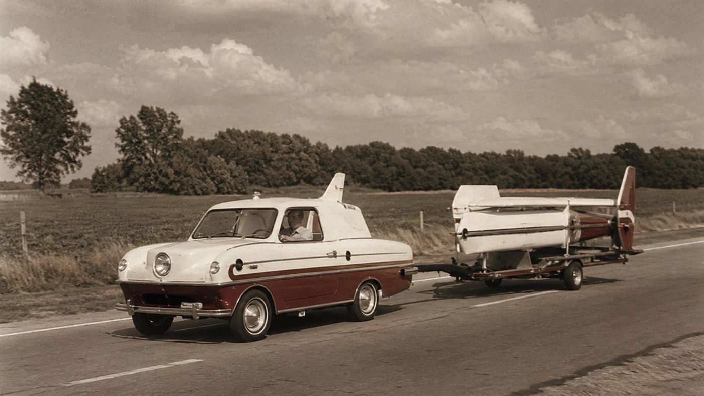 The Aerocar driving on a 1950s road as a compact car towing its folded wings behind it on a small trailer