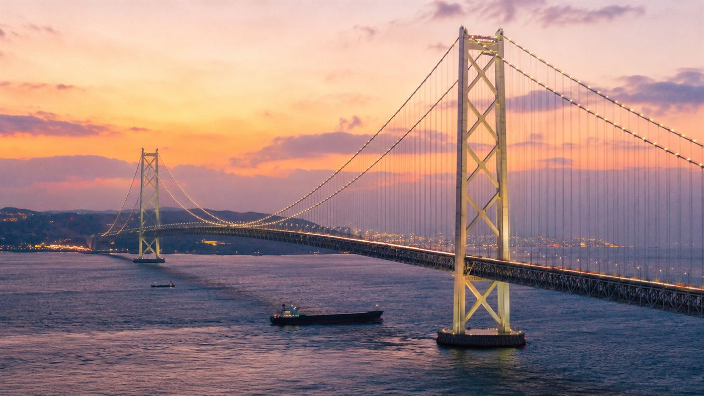 The Akashi Kaikyo Bridge, a vast white suspension bridge with two towers spanning a strait in Japan at dusk