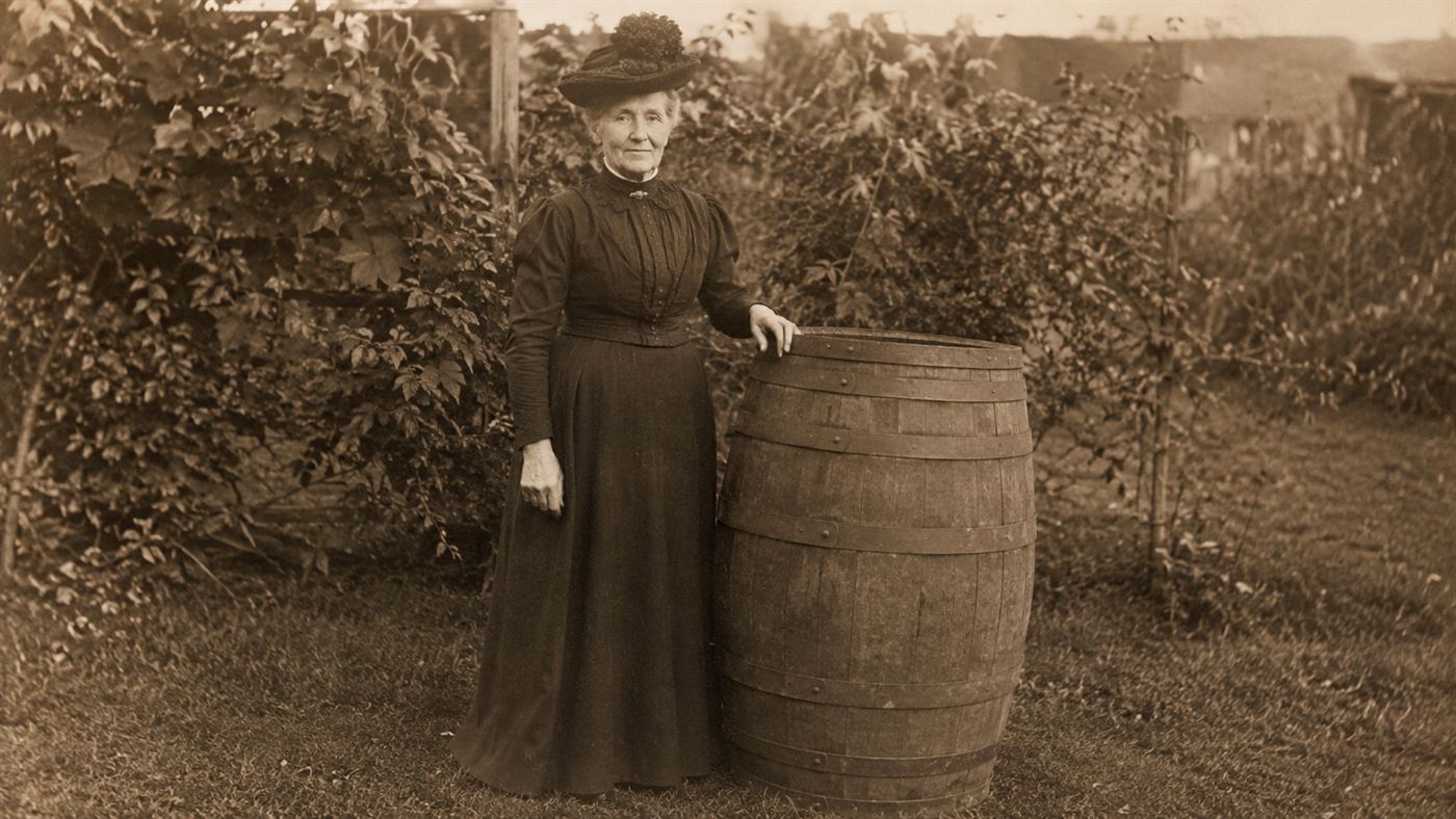 Annie Edson Taylor in Victorian dress standing beside the wooden barrel she rode over Niagara Falls in 1901