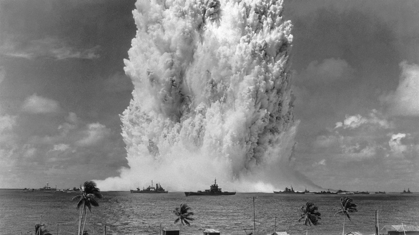 The Baker underwater nuclear test explosion at Bikini Atoll in 1946, an enormous white column of water and radioactive spray rising from the Pacific lagoon with navy warships visible below in the Marshall Islands