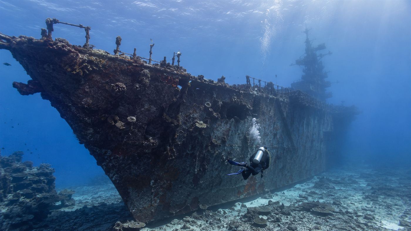 Scuba diver exploring the sunken USS Saratoga aircraft carrier wreck at Bikini Atoll in the Marshall Islands, coral and tropical fish covering the rusting hull in clear blue Pacific water, nuclear fallout legacy become diving site