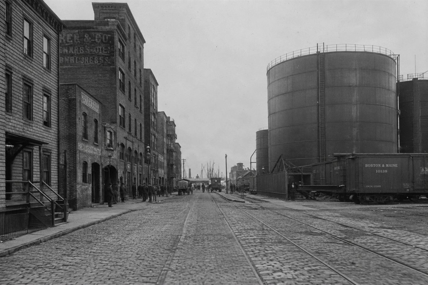 Boston Molasses Flood 1919 North End waterfront neighborhood, large cylindrical steel molasses storage tank looming over cobblestone street near harbor in winter