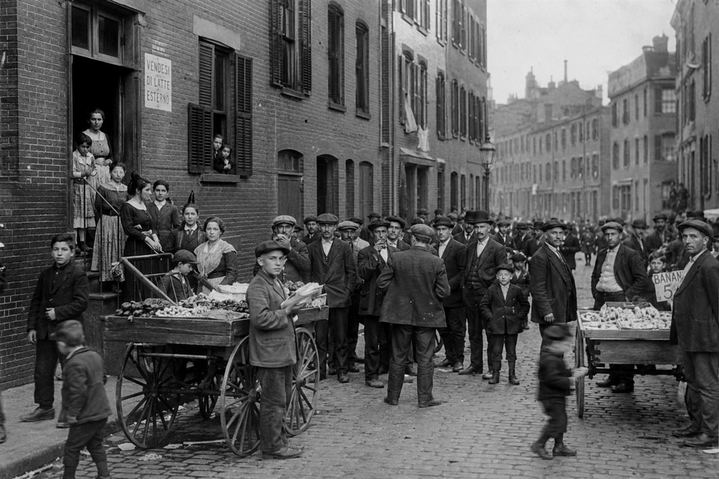 Italian immigrant workers and families outside red brick tenement buildings in Boston's North End around 1910s, period clothing, pushcarts on cobblestone street, grey winter light