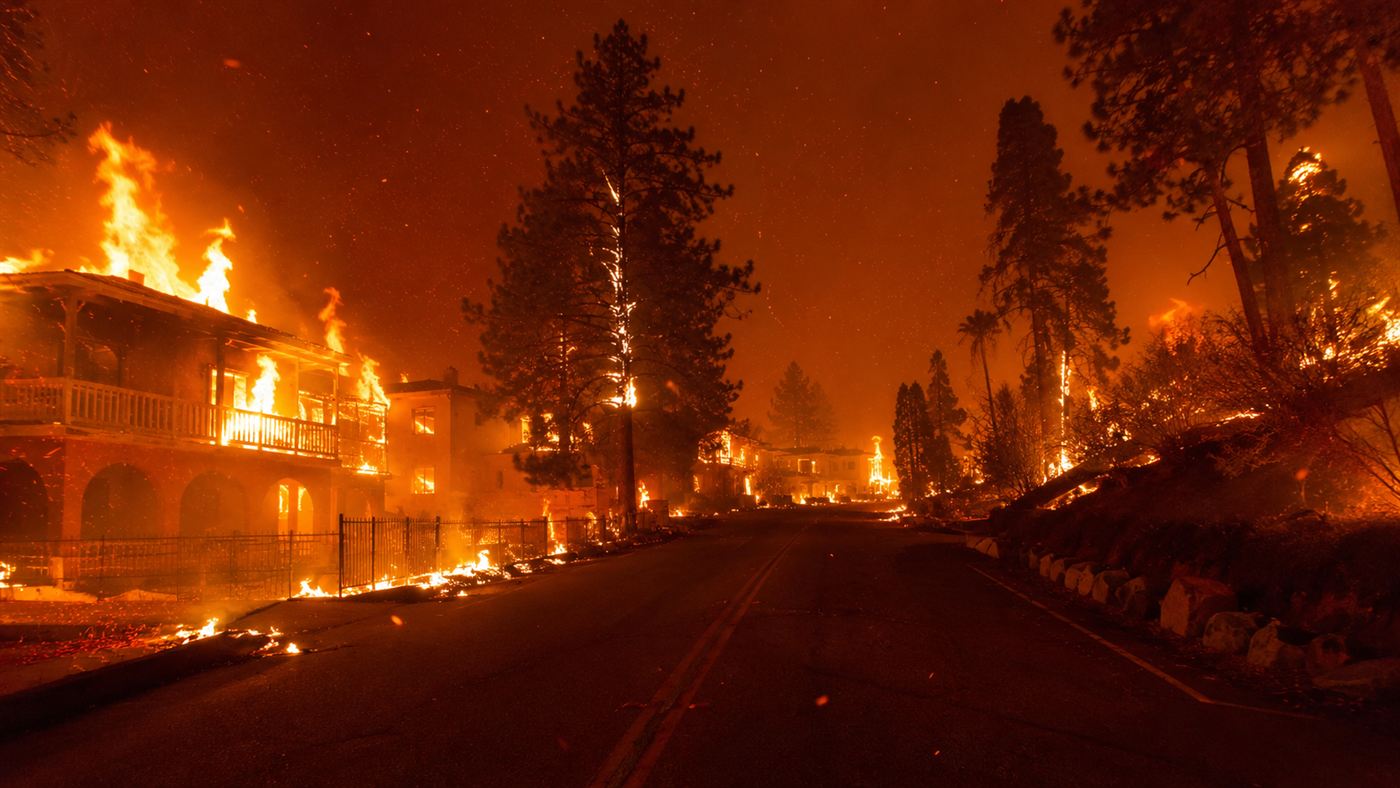 A California town engulfed by the Camp Fire under a dark orange smoke-filled sky, houses and pines burning as a road leads into the smoke