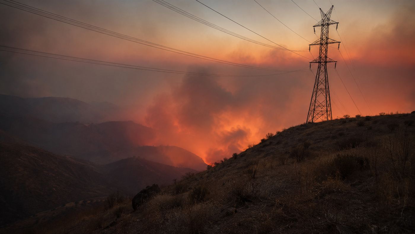 An old steel high-voltage power line transmission tower on a dry ridge with wildfire smoke and orange glow in the canyon below