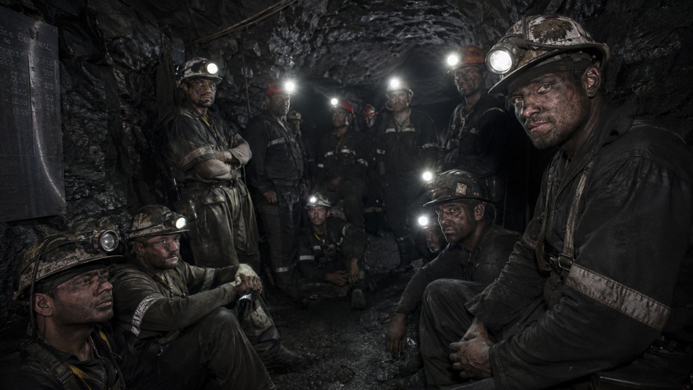 Exhausted dusty miners crowded in a dark underground refuge lit by helmet lamps