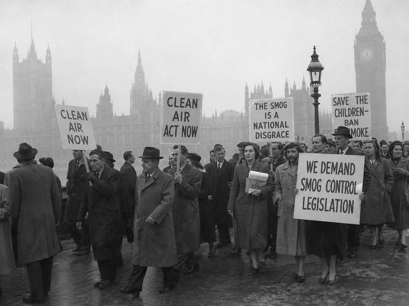 British protesters holding Clean Air Act banners outside the Houses of Parliament in the mid-1950s, demanding air pollution legislation after the Great Smog of 1952, smoggy urban backdrop, documentary style