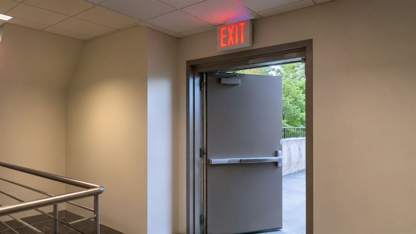 A modern illuminated exit sign over an outward-opening door with a panic push-bar, a fire safety code legacy
