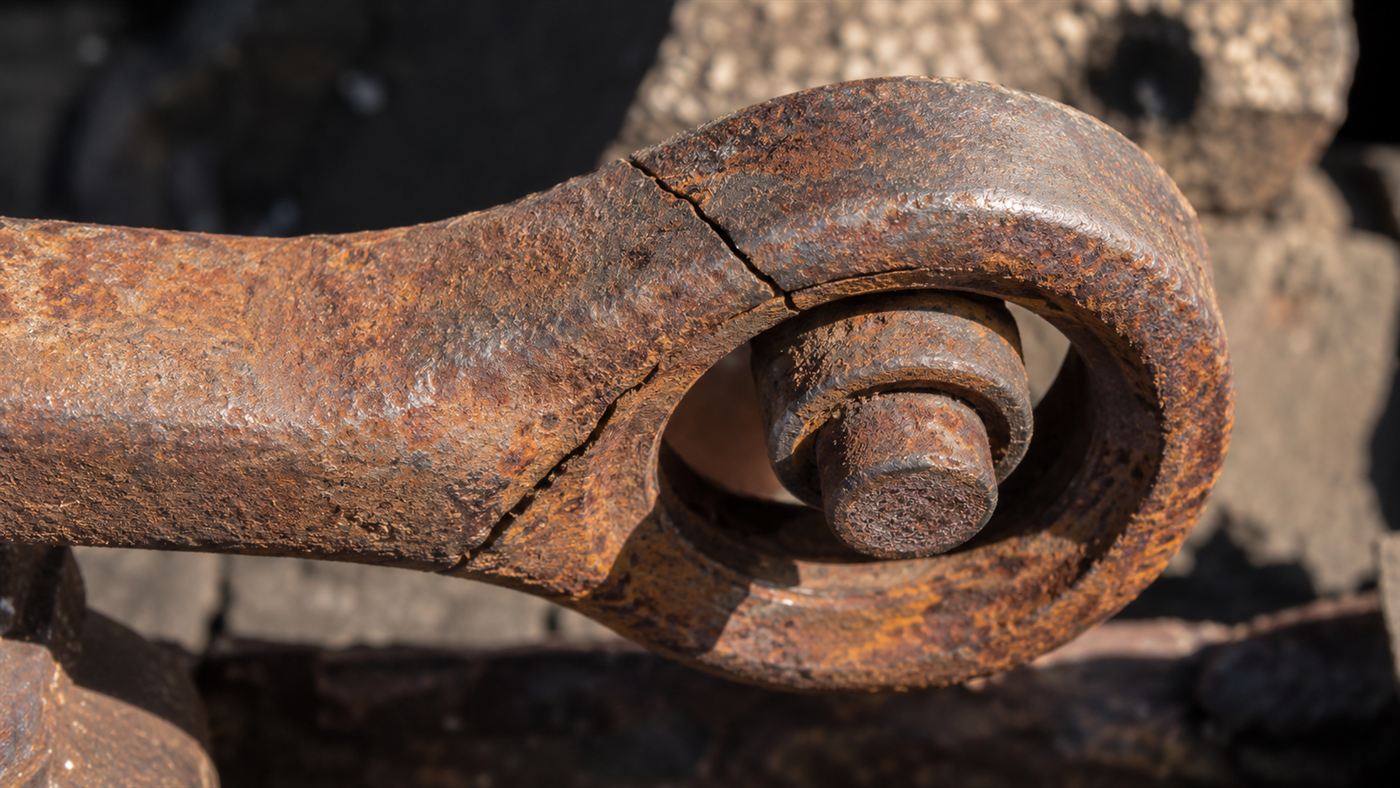 A close-up of a heavy rusted steel eyebar link with a pin joint and a fine crack running through the metal