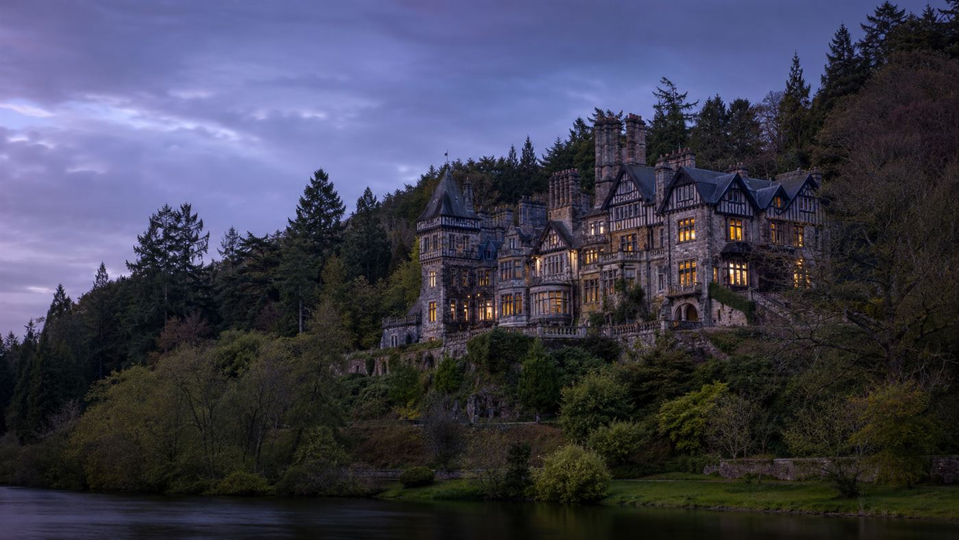 Cragside, a grand Victorian mansion on a wooded Northumberland hillside at dusk with a few windows glowing from early electric light
