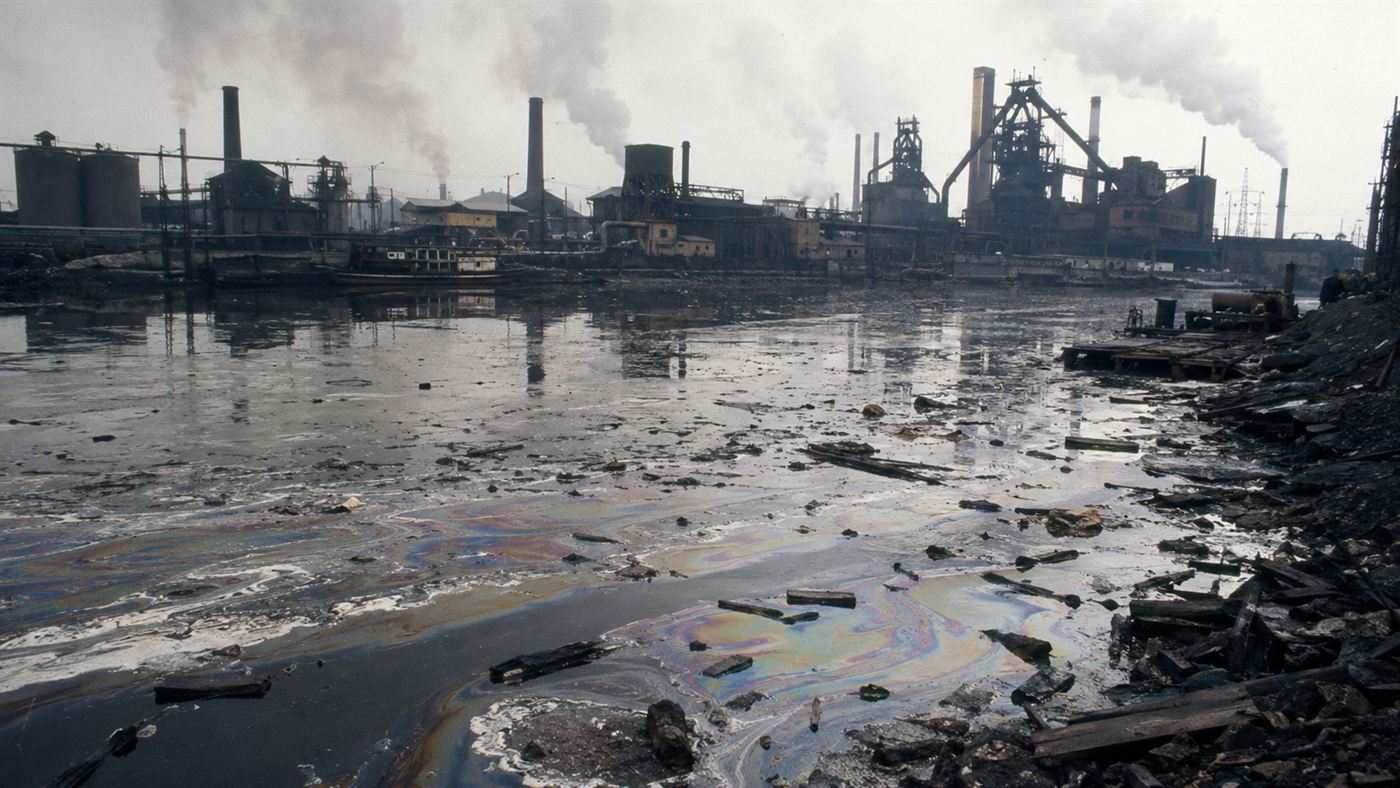 A sludgy, oil-slicked industrial river choked with foam and debris beside smoking factories