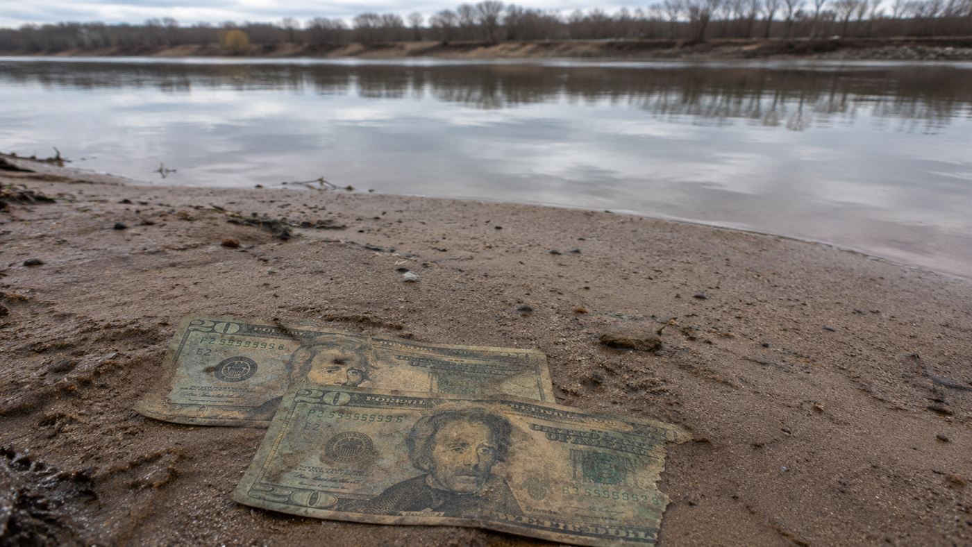 Weathered, decaying twenty-dollar bills partly buried in the sand of a river bank