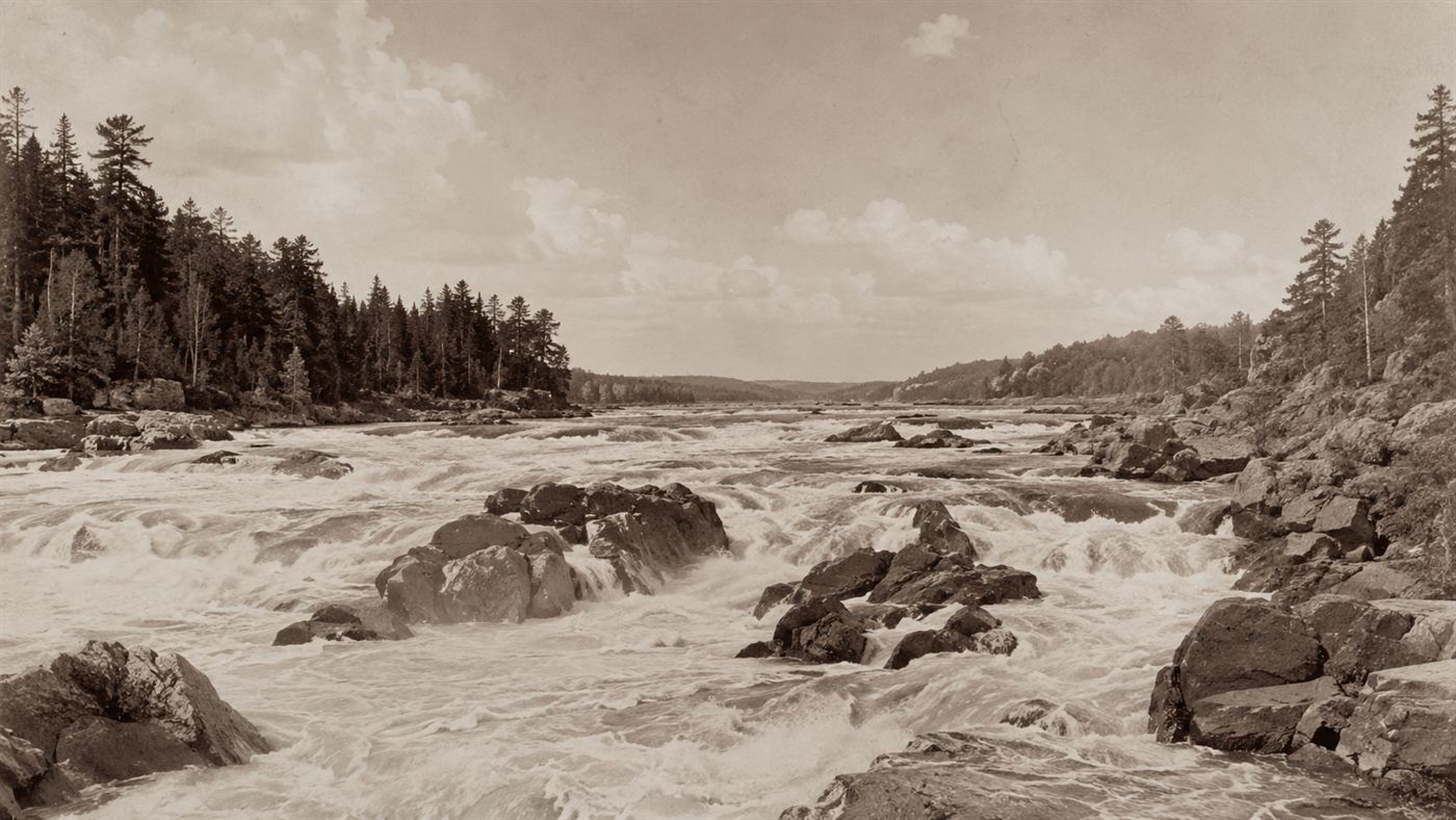 A wide stretch of the Mississippi River broken into fast rocky rapids and white water under a big sky, before it was dammed