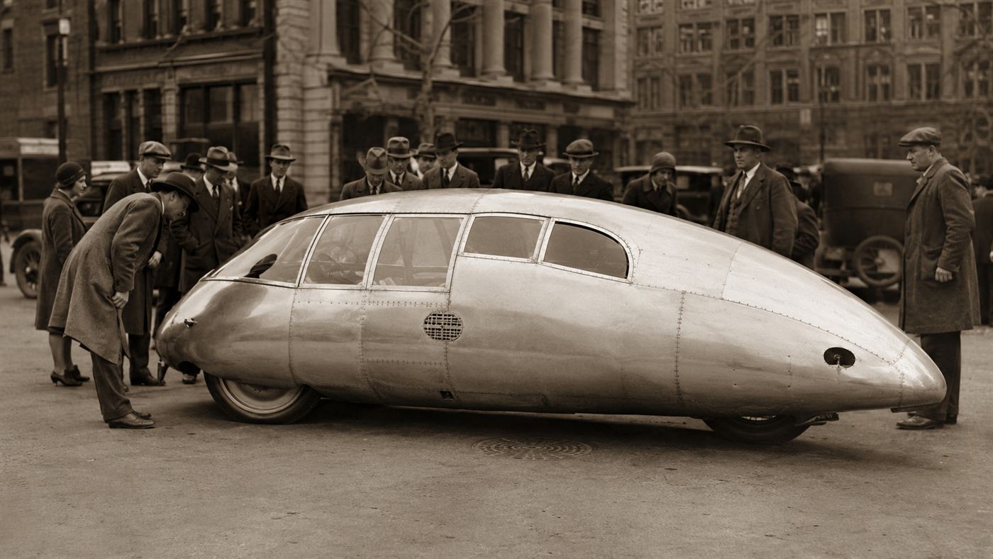 The 1933 Dymaxion car, a long silver teardrop-shaped aluminium vehicle on three wheels, parked on a street with onlookers