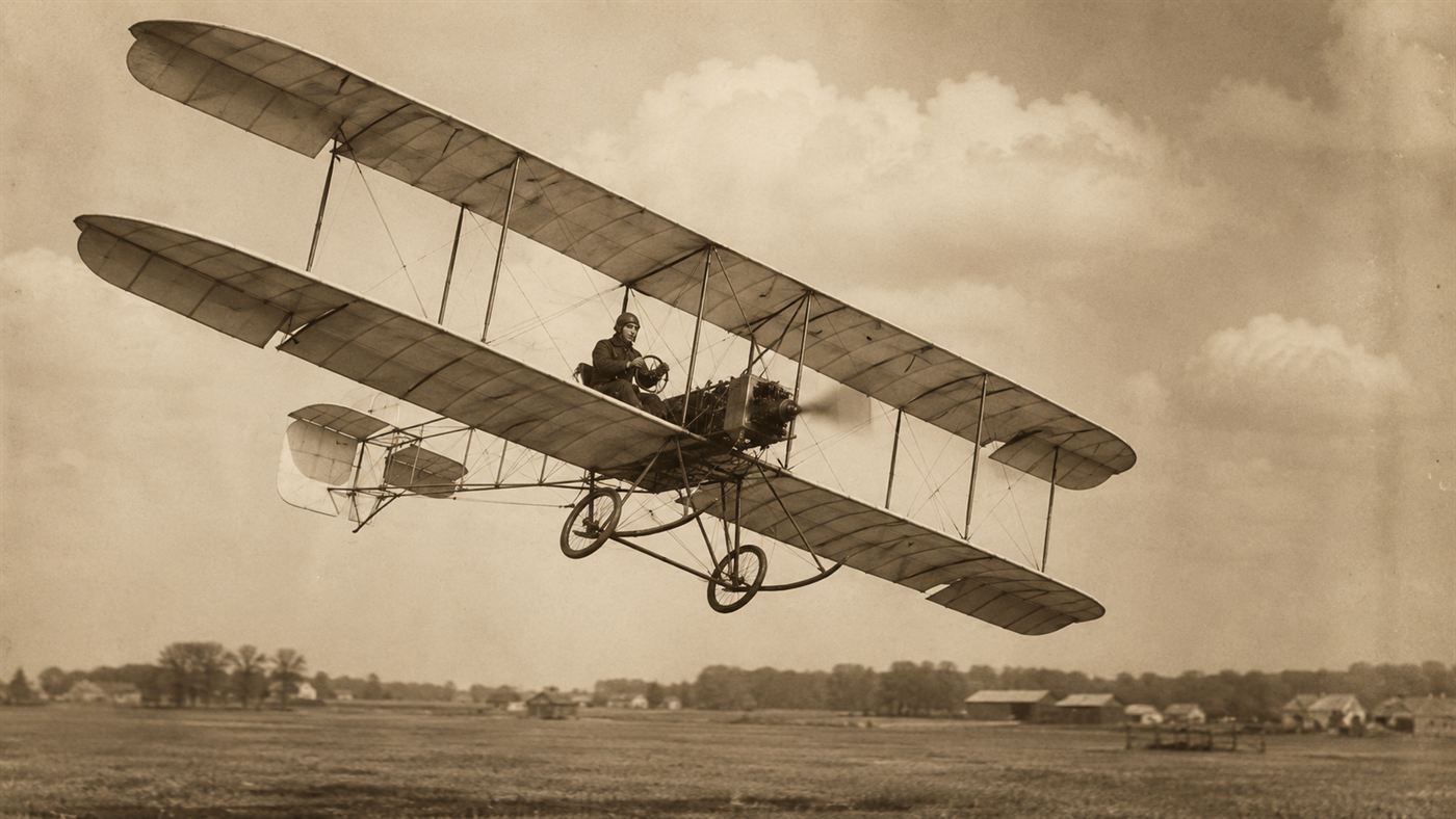 An early 1910s biplane with hinged ailerons banking in flight above a grass field, an aviator seated at the open controls