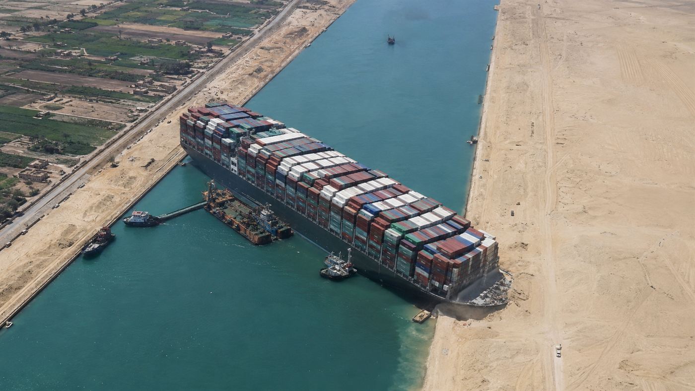 The Ever Given, an enormous container ship, wedged diagonally across a narrow desert canal, its bow buried in the bank, blocking all traffic