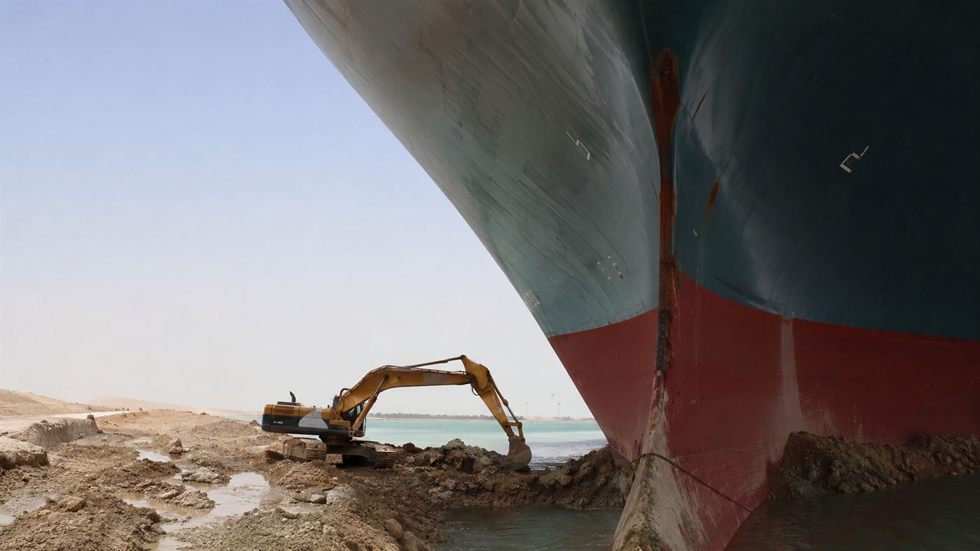 A small yellow excavator digging sand at the base of the towering bow of the beached Ever Given, part of the salvage operation