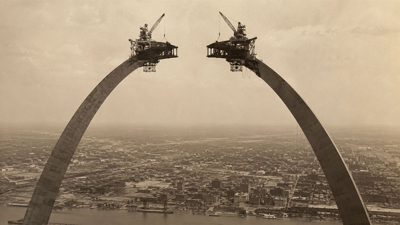 The two unfinished legs of the Gateway Arch under construction, curving up toward each other with creeper cranes